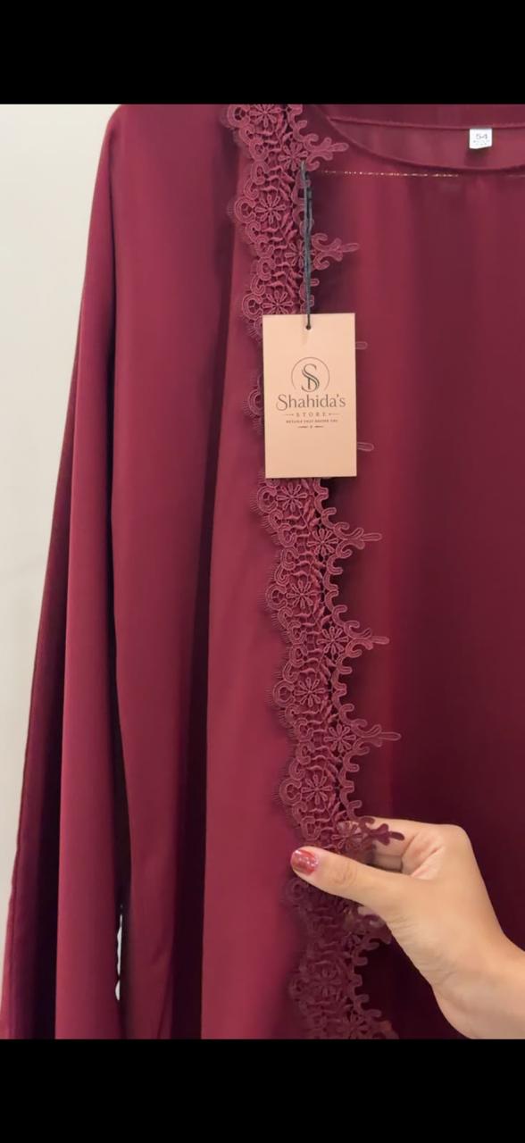 A close-up of a burgundy long-sleeve top with a matching lace trim down the front, featuring a hanging paper tag and a hand pointing to the lace detail.